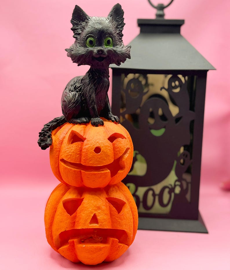 May include: A Halloween decoration featuring a black cat figurine with green eyes perched atop two stacked orange jack-o'-lanterns. A black lantern with ghost cutouts is in the background. The background is a solid pink color.