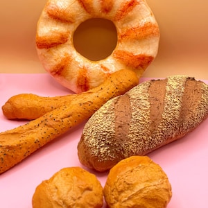May include: A selection of baked goods, including a large bagel with orange streaks, two baguettes, a loaf of bread, and two rolls. The bread products are various shades of brown and tan, set against a pink surface and a peach-coloured background.