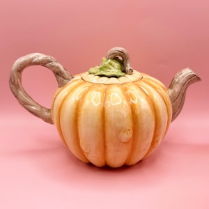 May include: A ceramic pumpkin-shaped teapot in light orange and cream. The handle and spout are designed to resemble tree branches. The lid has a green leaf and stem. The teapot is set against a pink background.