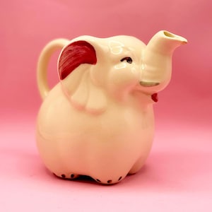 Puede incluir: Una jarra de cerámica blanca en forma de elefante con detalles pintados en rojo. La jarra tiene un pico y una asa.