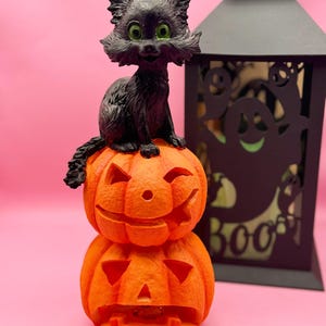 May include: A black cat figurine with green eyes sits atop two carved orange pumpkins. A black lantern with ghost cutouts is in the background. The pumpkins have carved faces, and the cat has a smiling expression. Halloween-themed decor.
