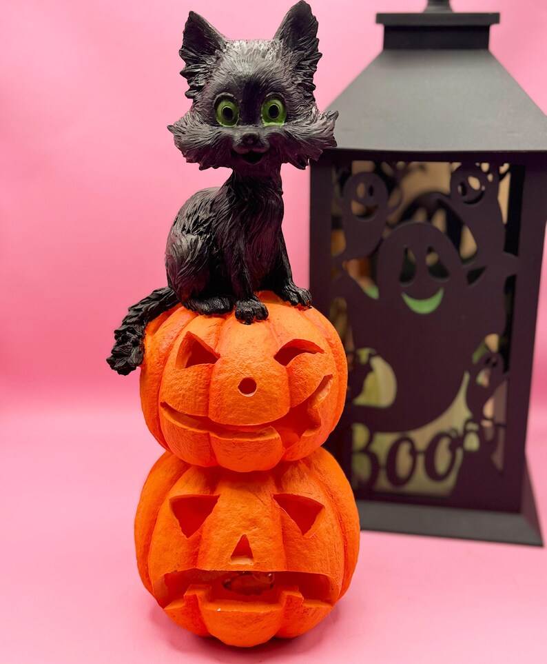 May include: A black cat figurine with green eyes sits atop two carved orange pumpkins. A black lantern with ghost cutouts is in the background. The pumpkins have carved faces, and the cat has a smiling expression. Halloween-themed decor.