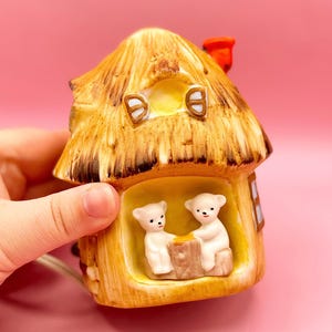 Puede incluir: Una luz de noche de cerámica en forma de cabaña con un techo de paja y dos figuritas de osos blancos en el interior. La cabaña está pintada en tonos marrones y amarillos, con una chimenea roja.
