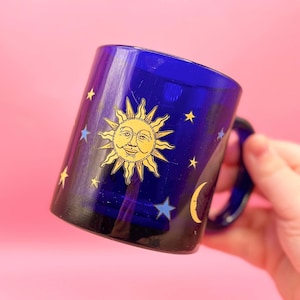 May include: A cobalt blue glass mug with a gold sun and moon design. The sun has a face and the moon is a crescent shape. There are gold stars scattered around the mug.