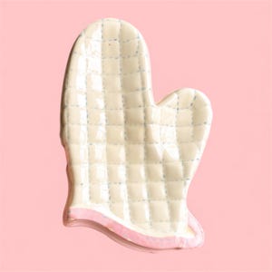 May include: Cream-coloured ceramic oven mitt with a quilted pattern and a pink trim. The mitt is designed with a thumb and a space for the fingers. The background is a solid pink.