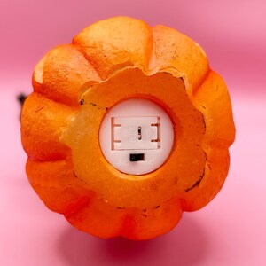 May include: Close-up of an orange pumpkin-shaped decoration with a textured surface. The bottom reveals a white compartment with an on/off switch. The pumpkin is set against a pink background, suggesting a Halloween or autumn theme.