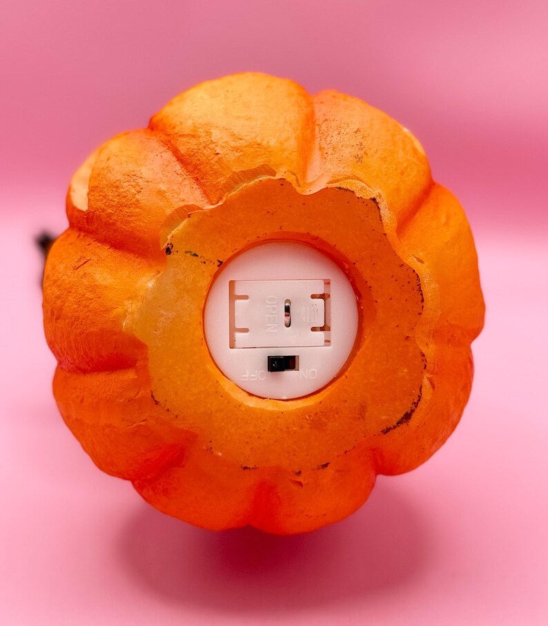 May include: Close-up of an orange pumpkin-shaped decoration with a textured surface. The bottom reveals a white compartment with an on/off switch. The pumpkin is set against a pink background, suggesting a Halloween or autumn theme.