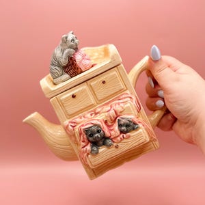 May include: A beige ceramic teapot with a cat and yarn basket on top. The teapot has a drawer design with two kittens peeking out from under a blanket. The handle and spout are curved.