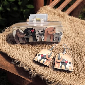 May include: A rectangular hair clip and matching earrings featuring an elephant design. The clip has a clear top and a beige base with elephant silhouettes in various colors. The earrings are wooden with elephant designs and silver hooks.