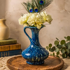 May include: A glossy blue ceramic pitcher vase with a speckled white and light green pattern, featuring a curved handle and a narrow neck. The vase is filled with white carnation flowers and two iridescent peacock feathers with blue eyes.