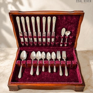 May include: A wooden flatware storage chest with a deep burgundy velvet interior lining, containing a set of silver-toned knives, spoons, and forks arranged in dedicated slots. The chest features a hinged lid and a logo reading COAL RIVER STUDIO.