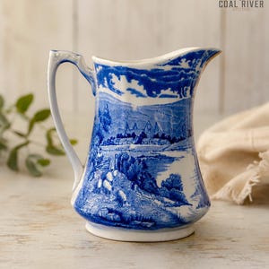 May include: A white ceramic pitcher featuring a blue transferware pattern depicting a landscape with rolling hills, trees, and buildings. The handle is curved, and the rim has a distinct pouring spout. The surface displays a detailed scene.