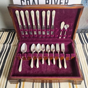 May include: A wooden storage chest with a deep purple velvet interior containing a set of silver-toned flatware. The collection includes eight knives, six spoons of varying sizes, two forks, and two small serving utensils arranged in dedicated slots.