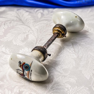 May include: A white porcelain door knob set featuring a vintage car illustration and the text "1901 OLDSMOBILE RUNABOUT" on one side. The set includes two oval knobs connected by a central metal spindle with dark, weathered brass-toned hardware.
