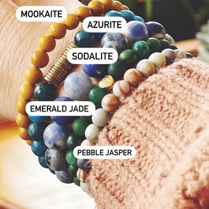 May include: A stack of beaded bracelets featuring various gemstones, including Mookaite, Azurite, Sodalite, Emerald Jade, and Pebble Jasper. The bracelets are in shades of yellow, blue, green, and white. A gold-colored spacer is visible.