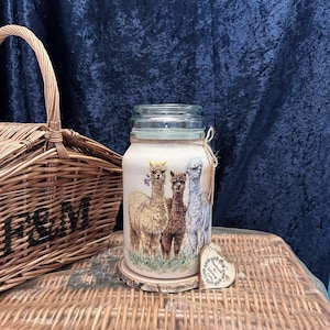 May include: A clear glass jar with a wide mouth and a screw-top lid, featuring a decorative wrap depicting three alpacas in cream, brown, and grey colours. A small wooden heart-shaped tag is tied to the neck with twine, resting on a wood slice.