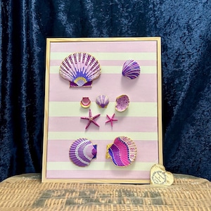 May include: A rectangular wooden frame containing a light pink and white striped canvas decorated with various hand-painted seashells and two starfish in shades of purple, pink, and white. A small heart-shaped tag with text is at the bottom right.