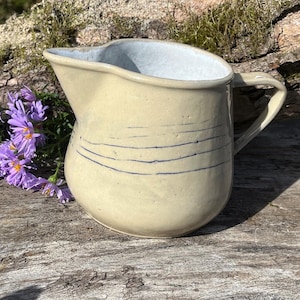 Pode incluir: Jarro de cerâmica artesanal com exterior creme e interior azul claro. O jarro apresenta uma pega curva e linhas azuis horizontais. É exibido numa superfície de madeira com flores roxas.