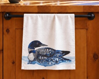 May include: A white kitchen towel hanging on a black metal bar, featuring a detailed illustration of a black and white loon swimming in blue water. The towel is rectangular and made of fabric, displaying the bird in a side profile view.