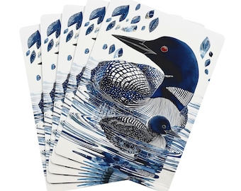 May include: A stack of playing cards featuring a watercolor illustration of a loon and its chick swimming in water. The cards have a white background with blue accents, including leaves and water details. The loons are depicted in shades of blue and black.