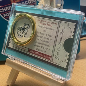May include: A genuine carousel brass ring from Gillian's Wonderland Pier, presented on a miniature wooden easel. The ring is in a clear acrylic case with a teal backing. The card reads "Grab the Brass Ring!"