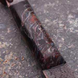 Peut inclure: Gros plan sur un manche de couteau personnalis&eacute; avec un motif en r&eacute;sine marbr&eacute;e dans les tons noir, marron, bleu et blanc. Le manche pr&eacute;sente des accents cuivr&eacute;s et une lame en m&eacute;tal fonc&eacute; textur&eacute;.