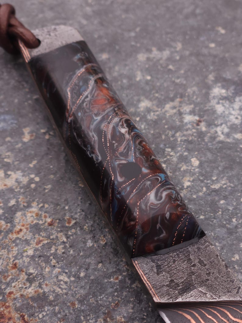 Peut inclure: Gros plan sur un manche de couteau personnalis&eacute; avec un motif en r&eacute;sine marbr&eacute;e dans les tons noir, marron, bleu et blanc. Le manche pr&eacute;sente des accents cuivr&eacute;s et une lame en m&eacute;tal fonc&eacute; textur&eacute;.