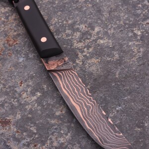 Peut inclure: Un couteau artisanal avec un manche noir et une lame cuivr&eacute;e. La lame pr&eacute;sente un motif unique de grain de bois. Deux rivets en cuivre fixent le manche. Une corde noire est attach&eacute;e au manche.