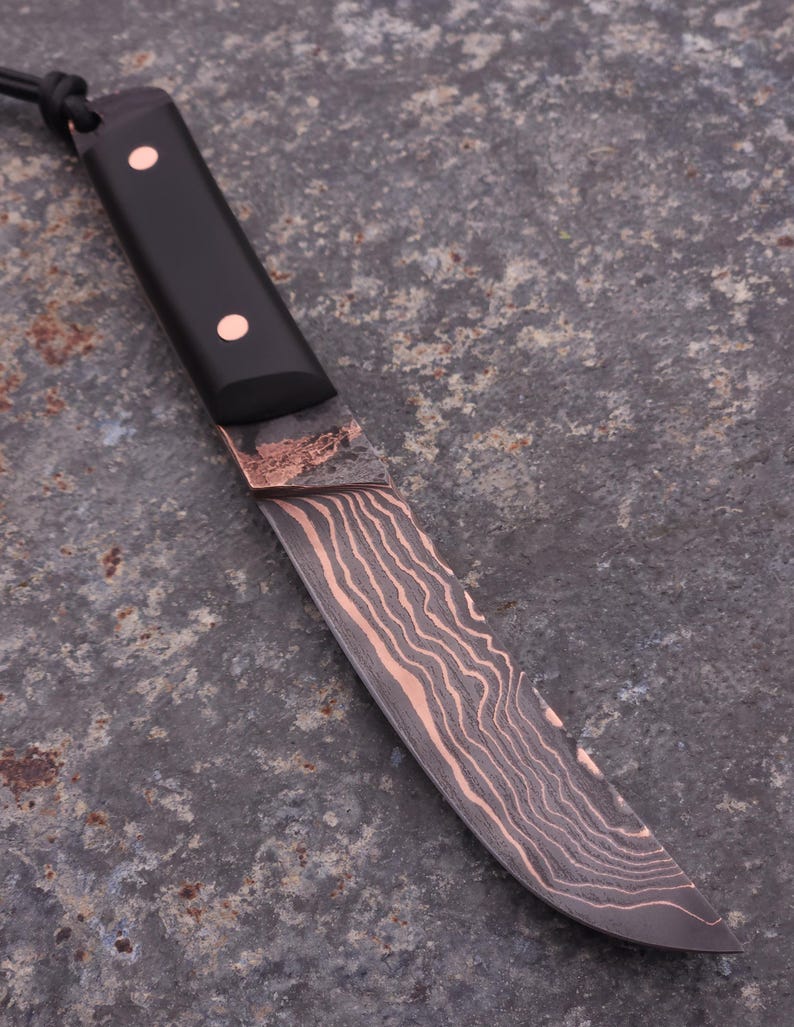 Peut inclure: Un couteau artisanal avec un manche noir et une lame cuivr&eacute;e. La lame pr&eacute;sente un motif unique de grain de bois. Deux rivets en cuivre fixent le manche. Une corde noire est attach&eacute;e au manche.