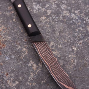 Peut inclure: Un couteau artisanal avec un manche sombre et une lame pr&eacute;sentant un motif unique de grain de bois. Le manche comporte deux petits accents ronds et clairs. Une lani&egrave;re en cuir est attach&eacute;e &agrave; l'extr&eacute;mit&eacute; du manche.
