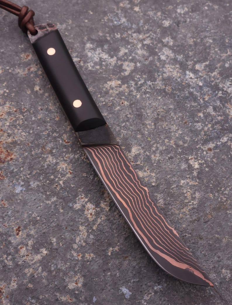 Peut inclure: Un couteau artisanal avec un manche sombre et une lame pr&eacute;sentant un motif unique de grain de bois. Le manche comporte deux petits accents ronds et clairs. Une lani&egrave;re en cuir est attach&eacute;e &agrave; l'extr&eacute;mit&eacute; du manche.