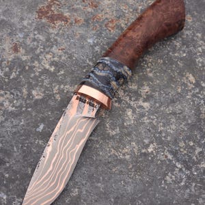 May include: A handcrafted knife with a curved blade and a wooden handle. The blade has a unique pattern and a copper bolster. The handle is made of dark wood with a burl pattern. The knife is designed for outdoor use.