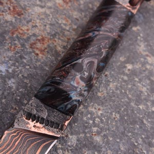 Peut inclure: Gros plan sur un couteau personnalis&eacute; avec une lame et un manche &agrave; motifs. La lame pr&eacute;sente un motif ondul&eacute; de couleur cuivr&eacute;e. Le manche pr&eacute;sente un motif marbr&eacute; dans les tons de noir, de bleu et de cuivre. Un cordon en cuir est attach&eacute; au manche.