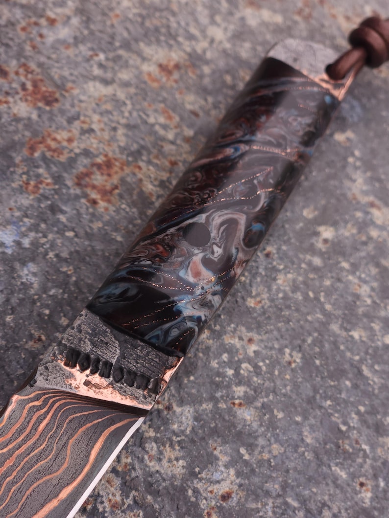 Peut inclure: Gros plan sur un couteau personnalis&eacute; avec une lame et un manche &agrave; motifs. La lame pr&eacute;sente un motif ondul&eacute; de couleur cuivr&eacute;e. Le manche pr&eacute;sente un motif marbr&eacute; dans les tons de noir, de bleu et de cuivre. Un cordon en cuir est attach&eacute; au manche.