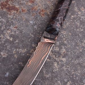 Peut inclure: Un couteau artisanal avec une lame &agrave; motifs et un manche sombre marbr&eacute;. La lame pr&eacute;sente un motif vein&eacute; de couleur cuivr&eacute;e. Le manche est sombre avec des tourbillons bleus et blancs. Un cordon en cuir est attach&eacute; &agrave; l'extr&eacute;mit&eacute; du manche.