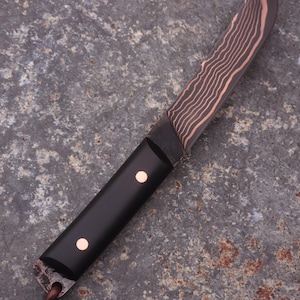 Peut inclure: Un couteau artisanal avec une lame sombre &agrave; motifs et un manche noir. La lame pr&eacute;sente un motif ondul&eacute; de couleur cuivr&eacute;e. Le manche comporte deux rivets de couleur cuivr&eacute;e. Une lani&egrave;re en cuir est attach&eacute;e &agrave; la base du manche.