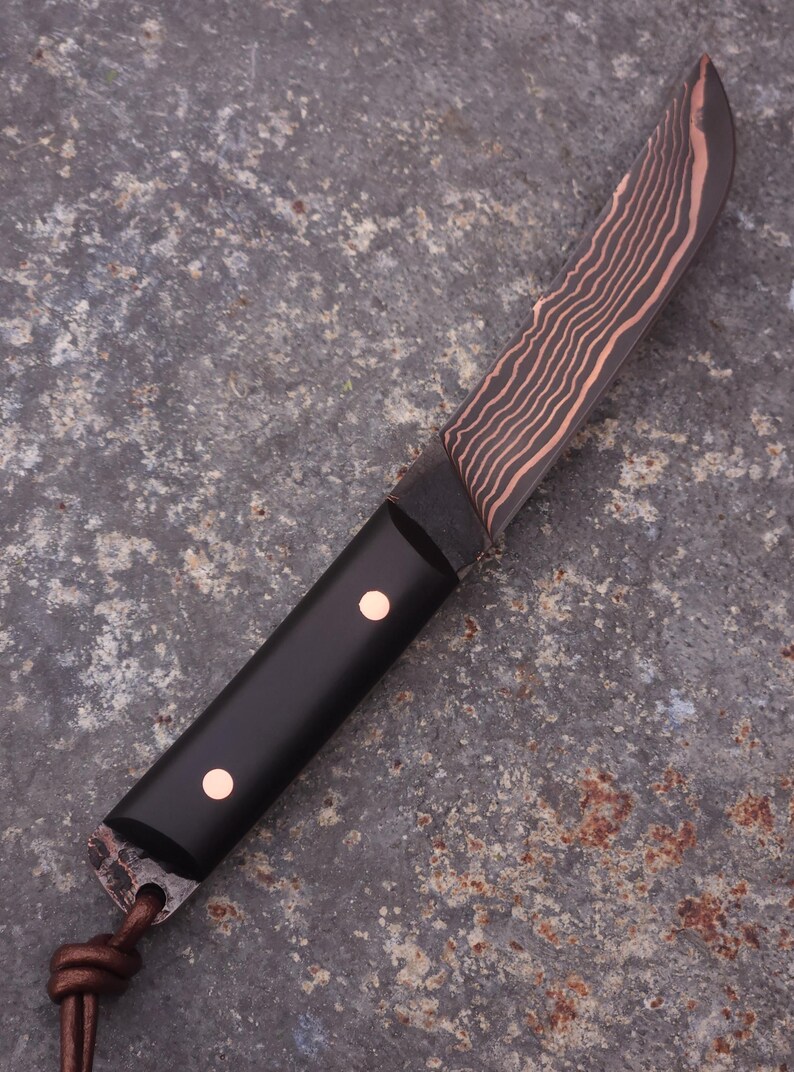 Peut inclure: Un couteau artisanal avec une lame sombre &agrave; motifs et un manche noir. La lame pr&eacute;sente un motif ondul&eacute; de couleur cuivr&eacute;e. Le manche comporte deux rivets de couleur cuivr&eacute;e. Une lani&egrave;re en cuir est attach&eacute;e &agrave; la base du manche.