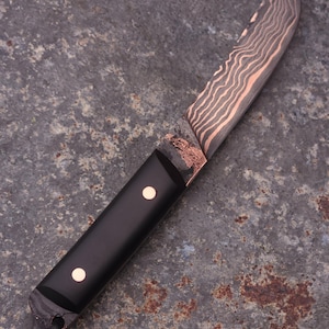 Peut inclure: Un couteau artisanal avec une lame &agrave; motifs et un manche noir. La lame pr&eacute;sente un motif de couleur cuivr&eacute;e, et le manche comporte deux petits accents ronds de couleur cuivr&eacute;e. Une lani&egrave;re en cuir est attach&eacute;e &agrave; la base du manche.