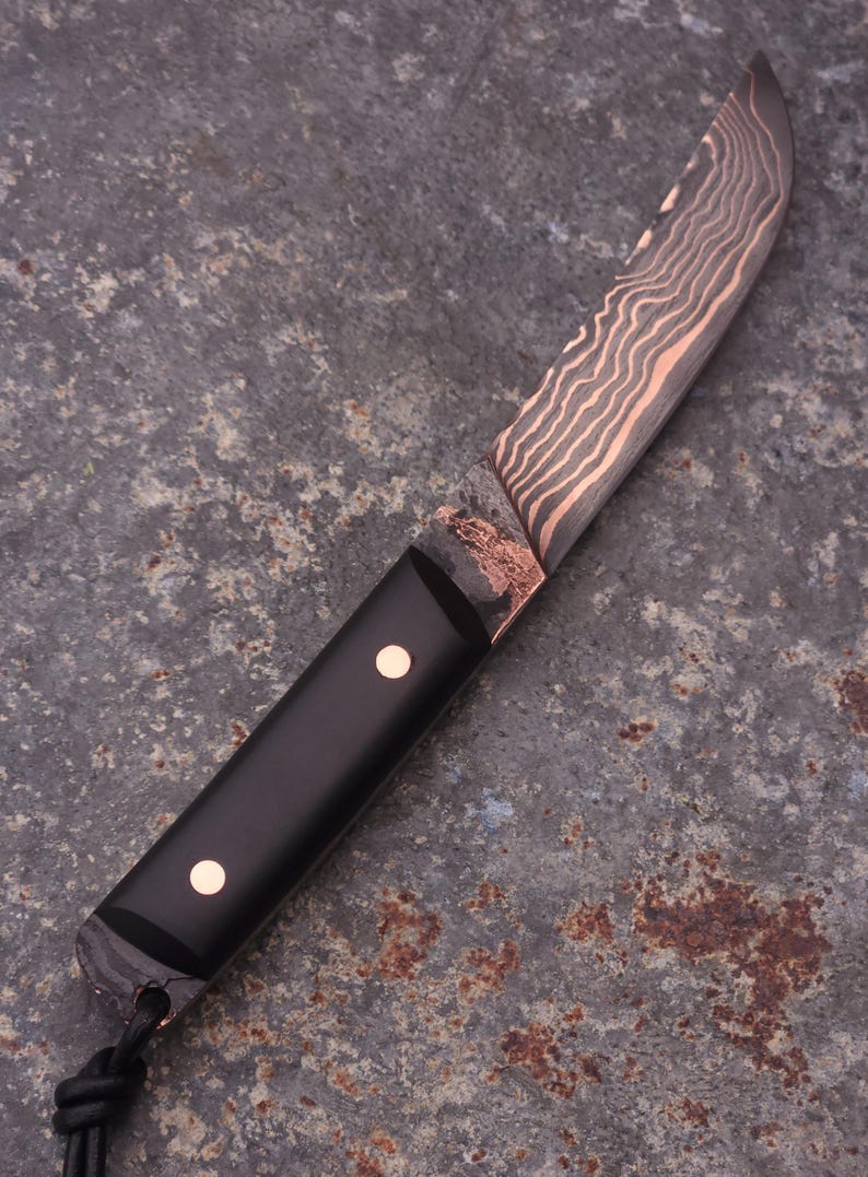 Peut inclure: Un couteau artisanal avec une lame &agrave; motifs et un manche noir. La lame pr&eacute;sente un motif de couleur cuivr&eacute;e, et le manche comporte deux petits accents ronds de couleur cuivr&eacute;e. Une lani&egrave;re en cuir est attach&eacute;e &agrave; la base du manche.