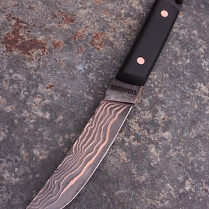 Peut inclure: Couteau artisanal avec un manche noir et des rivets en cuivre. La lame pr&eacute;sente un motif ondul&eacute; unique en cuivre et gris. Le mot "THORSHOL" est grav&eacute; pr&egrave;s du manche. Un cordon noir est attach&eacute; au manche.