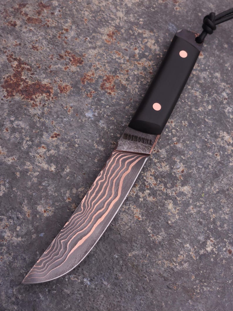 Peut inclure: Couteau artisanal avec un manche noir et des rivets en cuivre. La lame pr&eacute;sente un motif ondul&eacute; unique en cuivre et gris. Le mot "THORSHOL" est grav&eacute; pr&egrave;s du manche. Un cordon noir est attach&eacute; au manche.