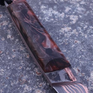Peut inclure: Gros plan sur un couteau artisanal. La lame pr&eacute;sente un motif distinctif, tandis que le manche est fait de r&eacute;sine marron fonc&eacute;, rouge et transparente avec des accents de cuivre. Une corde noire est attach&eacute;e au manche. La lame a un motif ondul&eacute;.