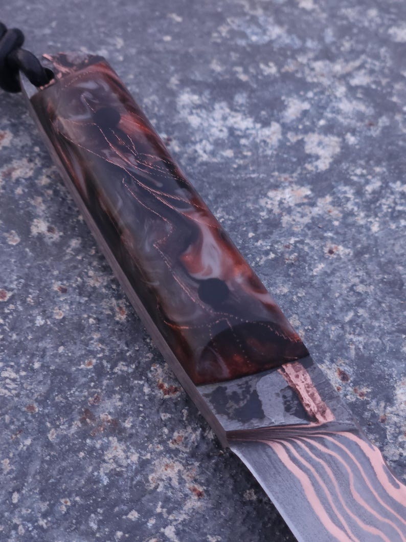 Peut inclure: Gros plan sur un couteau artisanal. La lame pr&eacute;sente un motif distinctif, tandis que le manche est fait de r&eacute;sine marron fonc&eacute;, rouge et transparente avec des accents de cuivre. Une corde noire est attach&eacute;e au manche. La lame a un motif ondul&eacute;.