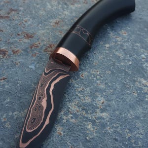 May include: A handcrafted knife with a black handle and a blade featuring a unique pattern of copper and dark metal. The knife has a copper bolster and decorative accents near the handle. The blade's design resembles wood grain.