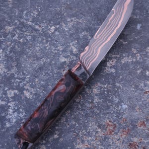 Peut inclure: Un couteau artisanal avec une lame &agrave; motifs et un manche sombre et marbr&eacute;. La lame pr&eacute;sente un motif cuivr&eacute; et le manche des nuances de rouge, de marron et de noir. Un cordon noir est attach&eacute; &agrave; la base du manche.