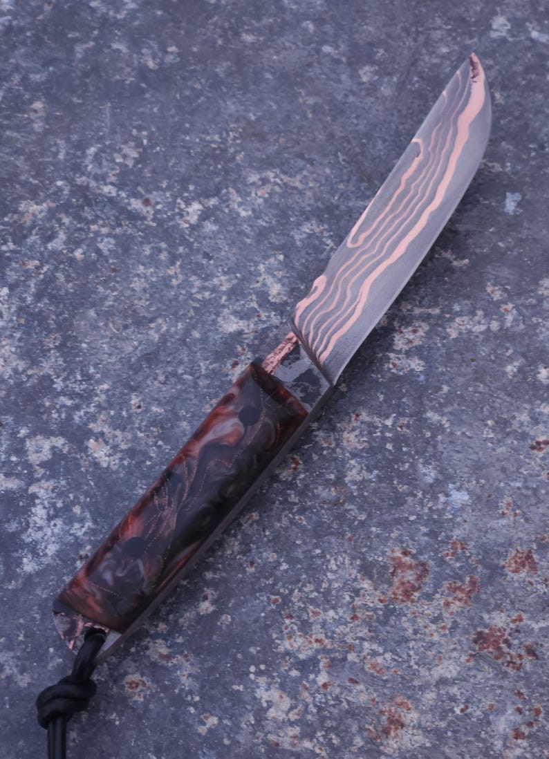 Peut inclure: Un couteau artisanal avec une lame &agrave; motifs et un manche sombre et marbr&eacute;. La lame pr&eacute;sente un motif cuivr&eacute; et le manche des nuances de rouge, de marron et de noir. Un cordon noir est attach&eacute; &agrave; la base du manche.