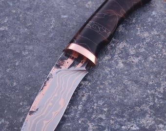 Peut inclure: Un couteau à lame fixe doté d'une lame en acier à motifs avec des lignes ondulées couleur cuivre et un manche en résine marbrée sombre. Le couteau comprend une mitre en cuivre poli séparant la lame du manche, avec du texte visible.