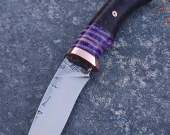 Peut inclure: Un couteau à lame fixe doté d'une lame en acier texturé avec un motif en couches visible. Le manche se compose d'une section en bois sombre, d'une entretoise rayée violette et blanche, et d'une mitre en cuivre poli avec une goupille.