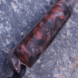 Peut inclure: Gros plan sur un manche de couteau avec un motif marbr&eacute; dans les tons de marron, blanc et noir. La poign&eacute;e est enferm&eacute;e dans un mat&eacute;riau transparent, avec des accents cuivr&eacute;s et un cordon en cuir noir. Le couteau semble fait &agrave; la main.