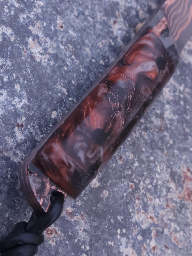 Peut inclure: Gros plan sur un manche de couteau avec un motif marbr&eacute; dans les tons de marron, blanc et noir. La poign&eacute;e est enferm&eacute;e dans un mat&eacute;riau transparent, avec des accents cuivr&eacute;s et un cordon en cuir noir. Le couteau semble fait &agrave; la main.